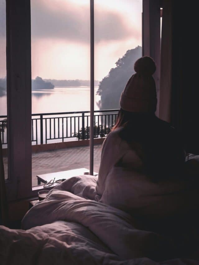Person wearing a beanie sitting by window, looking at a serene lake view in Pokhara.